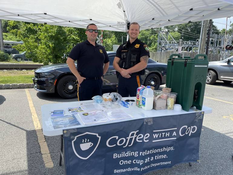 Two police officers posing at a table with a tank of coffee and cups. A banner on the table reads: Coffee with a cop. Building relationships. One cup at a time. Bernardsville Police Department.